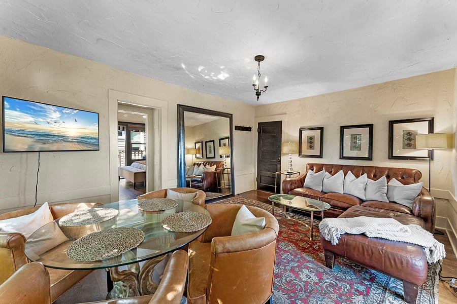 View from living room looking into bedroom and front door: Smart TV, wood blinds, oversized floor mirror, comfy leather love seat sectional with cushy pillows and soft cable knit blanket. Two side tables with lamps. Glass top coffee table. Glass top dining table with 4 chairs.
