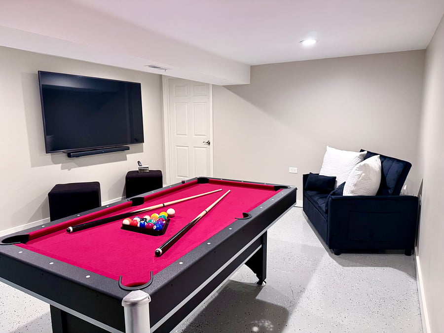 Multi-use entertainment room with 2 in 1 red felt pool/ping-pong table, large flatscreen TV and lounge area with barstool seating. Includes a couch that converts into a bed for additional sleeping space. Designed for fun, relaxation, and extra comfort.