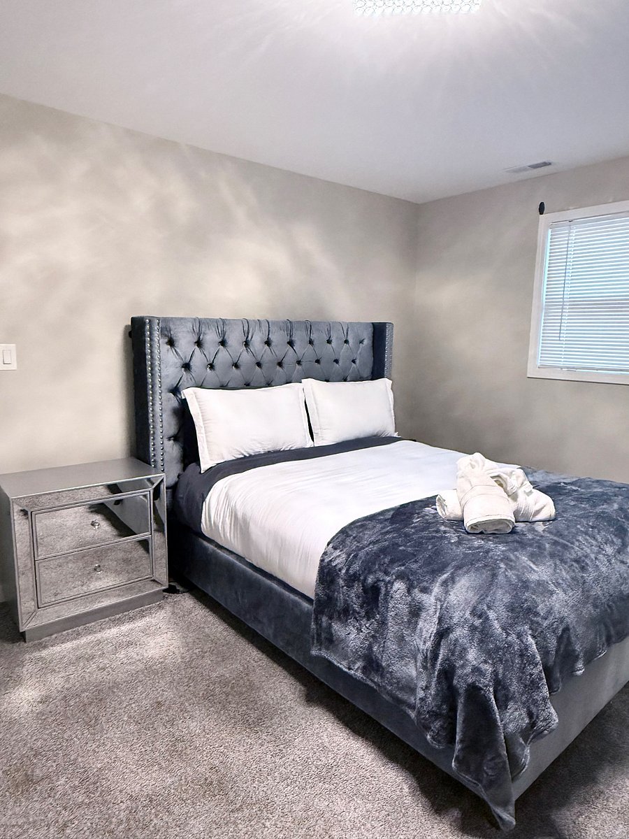Spacious bedroom with queen bed, upholstered tufted headboard & Wall mounted, Smart TV. Matching mirror, dresser, and nightstand set, seating bench, and a full body mirror for getting ready. Carpet flooring and window blinds for comfort & privacy.