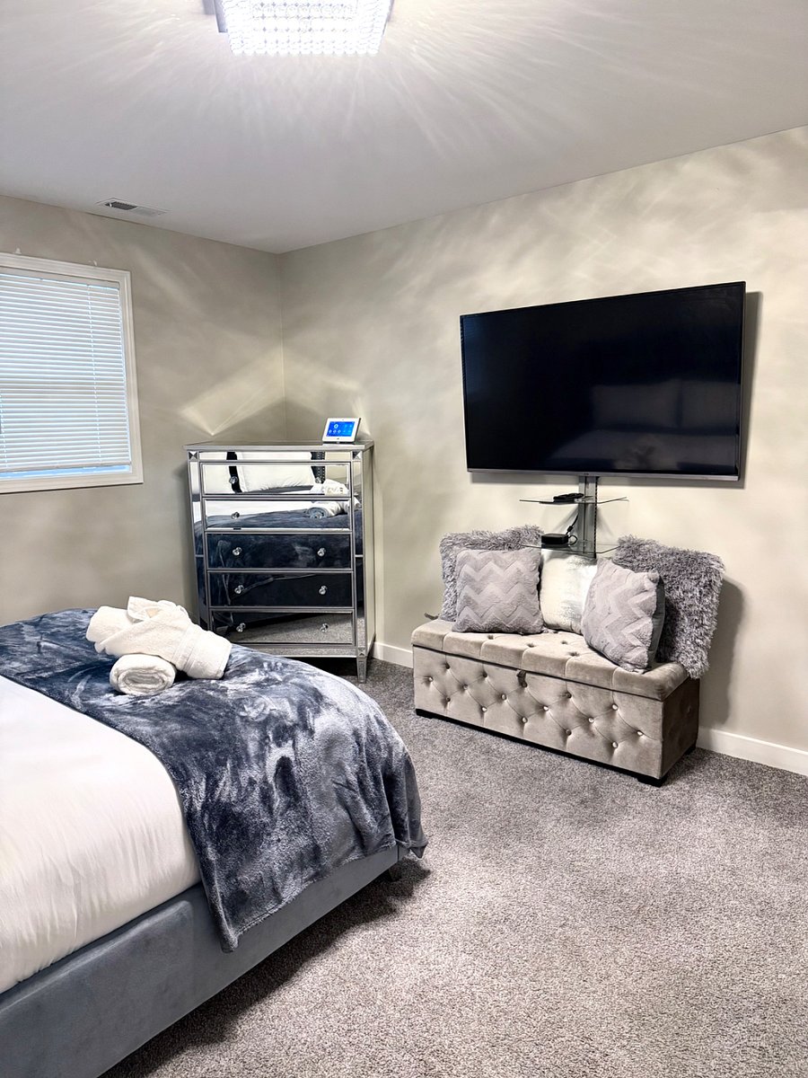 Spacious bedroom with queen bed, upholstered tufted headboard & Wall mounted, Smart TV. Matching mirror, dresser, and nightstand set, seating bench, and a full body mirror for getting ready. Carpet flooring and window blinds for comfort & privacy.