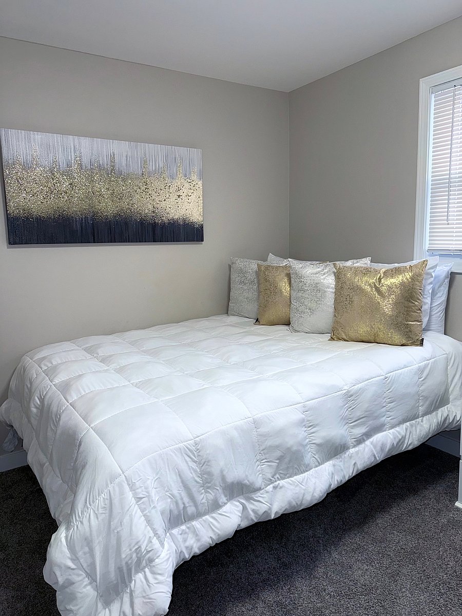 Cozy bedroom with the queen bed, dressed and soft, white bedding and decorative pillows. Features a night stand, wall art, a flatscreen TV and a desk with a comfort chair for work or study. Natural light from two windows with the blinds for brightness and privacy.