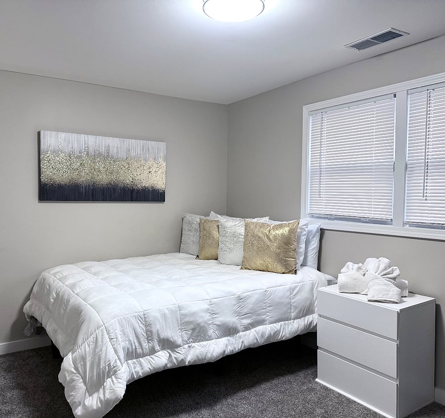 Cozy bedroom with the queen bed, dressed and soft, white bedding and decorative pillows. Features a night stand, wall art, a flatscreen TV and a desk with a comfort chair for work or study. Natural light from two windows with the blinds for brightness and privacy.