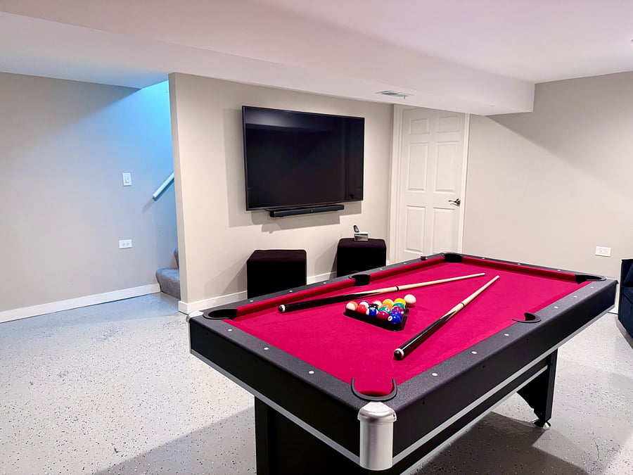 Multi-use entertainment room with 2 in 1 red felt pool/ping-pong table, large flatscreen TV and lounge area with barstool seating. Includes a couch that converts into a bed for additional sleeping space. Designed for fun, relaxation, and extra comfort.