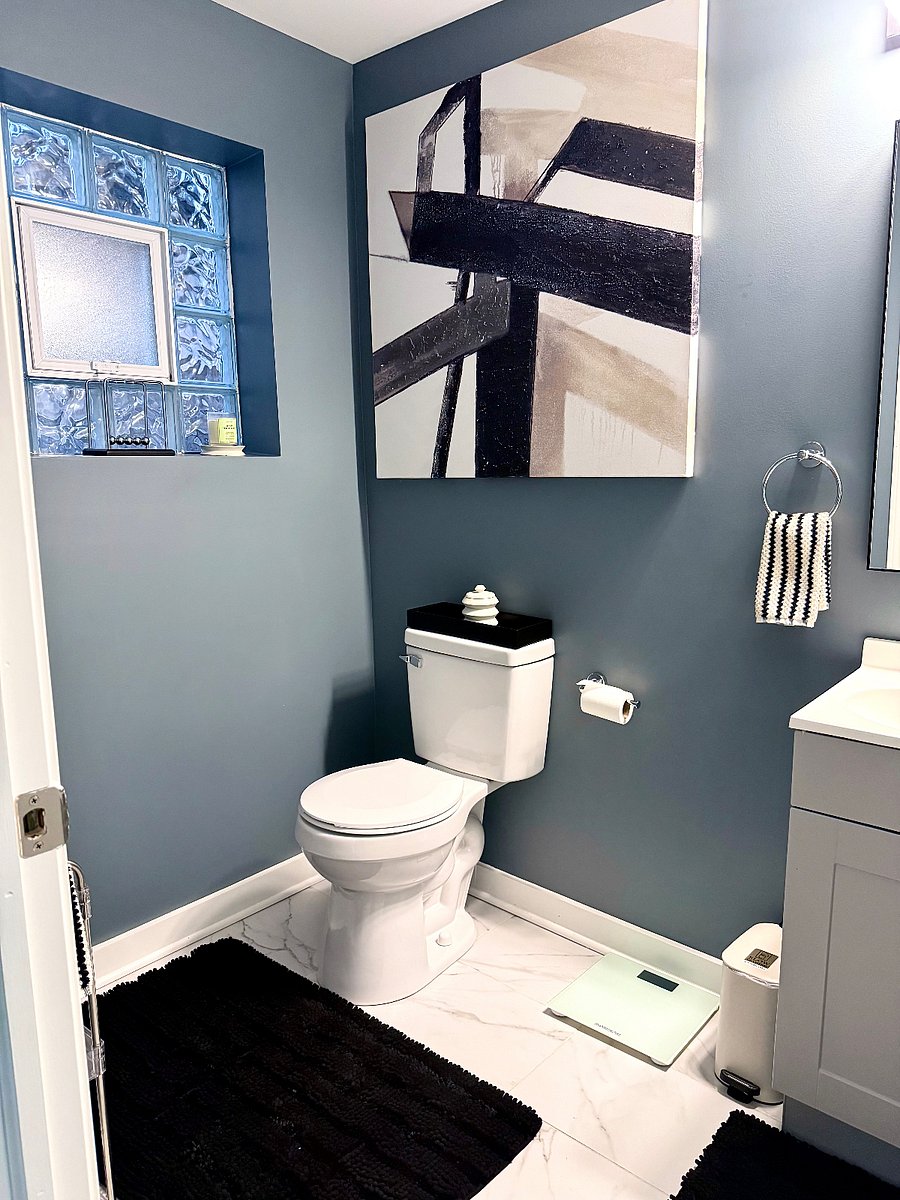 Decorated half bathroom with modern toilet, sink, vanity, Decorative hand towels in bold wall art. A shelving unit stocked with extra tissue, paper towels, and air freshener for guest convenience. A digital scale is also available for guest use.