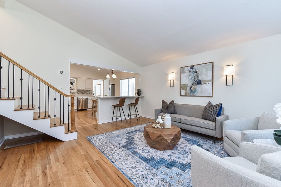 A stylish open-concept living room features modern furniture, warm wood flooring, and a seamless flow into a bright kitchen and dining area.
