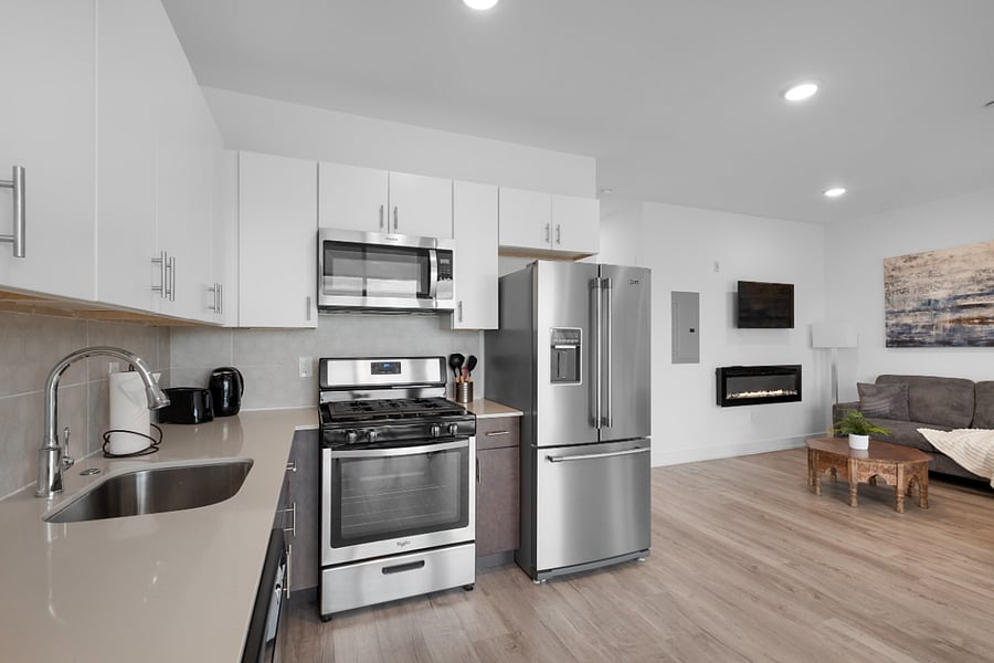 Bright, modern kitchen and living area with sleek stainless steel appliances, a stylish island with a marble countertop, and large windows that flood the space with natural light. The clean design and warm hardwood floors create a cozy yet contemporary vibe.