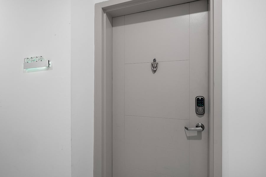 Sleek, modern white door set in a minimalist white wall. The door is equipped with a digital keypad lock and a peephole, emphasizing security and convenience.