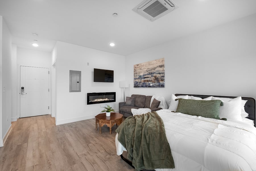 Cozy, modern studio apartment with a stylish mix of textures and colors. It features a neatly made bed near a large window, a patterned accent wall, and a seating area with a green loveseat and a navy blue chair around a hexagonal coffee table. Warm wood flooring and soft lighting create a welcoming, contemporary atmosphere.