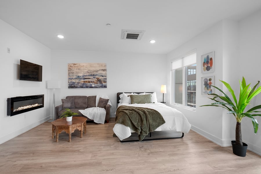 Cozy, modern studio apartment with a stylish mix of textures and colors. It features a neatly made bed near a large window, a patterned accent wall, and a seating area with a green loveseat and a navy blue chair around a hexagonal coffee table. Warm wood flooring and soft lighting create a welcoming, contemporary atmosphere.
