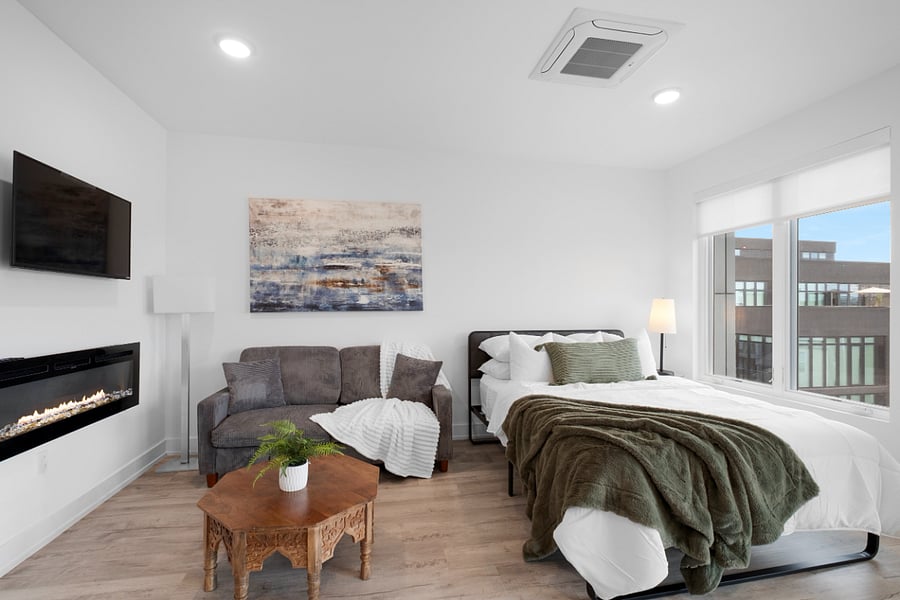 Cozy, modern studio apartment with a stylish mix of textures and colors. It features a neatly made bed near a large window, a patterned accent wall, and a seating area with a green loveseat and a navy blue chair around a hexagonal coffee table. Warm wood flooring and soft lighting create a welcoming, contemporary atmosphere.