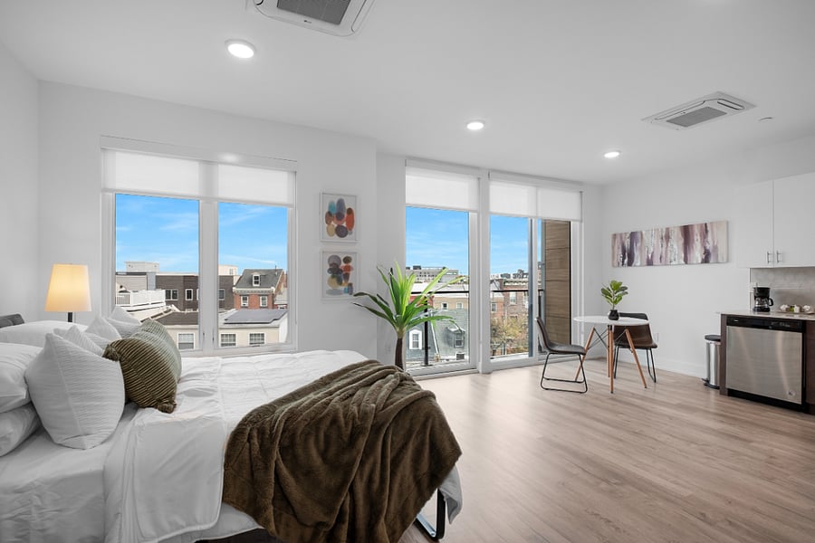 Bright, modern studio apartment with an open layout that combines kitchen, living, and bedroom areas. It features sleek cabinetry, stainless steel appliances, a central island with a sink, and warm hardwood floors. Large windows and a glass door lead to a balcony, filling the space with natural light and offering a view of nearby buildings.