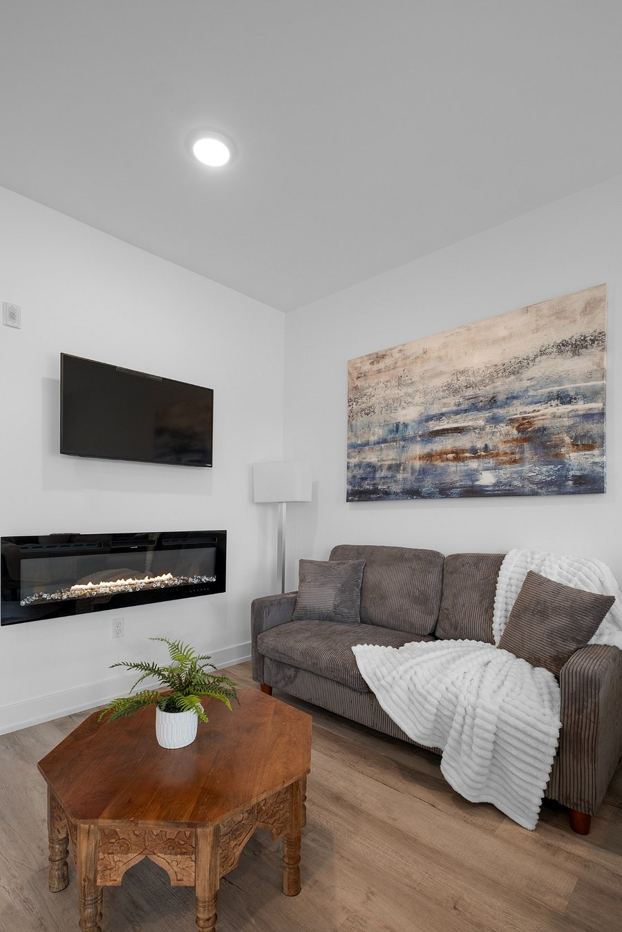 A cozy and modern living room corner featuring a gray upholstered sofa with a white textured throw, a carved wooden coffee table topped with a potted fern, and a sleek electric fireplace beneath a wall-mounted TV.