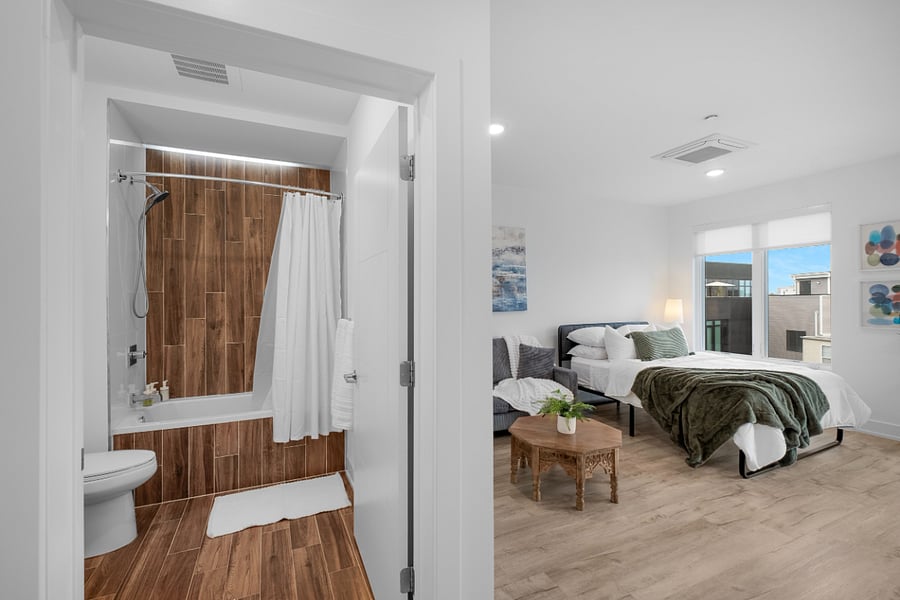 A bright, modern apartment interior featuring a bathroom and adjacent bedroom. The bathroom has warm wood-look tile, a white bathtub with a shower curtain, and a sleek toilet.