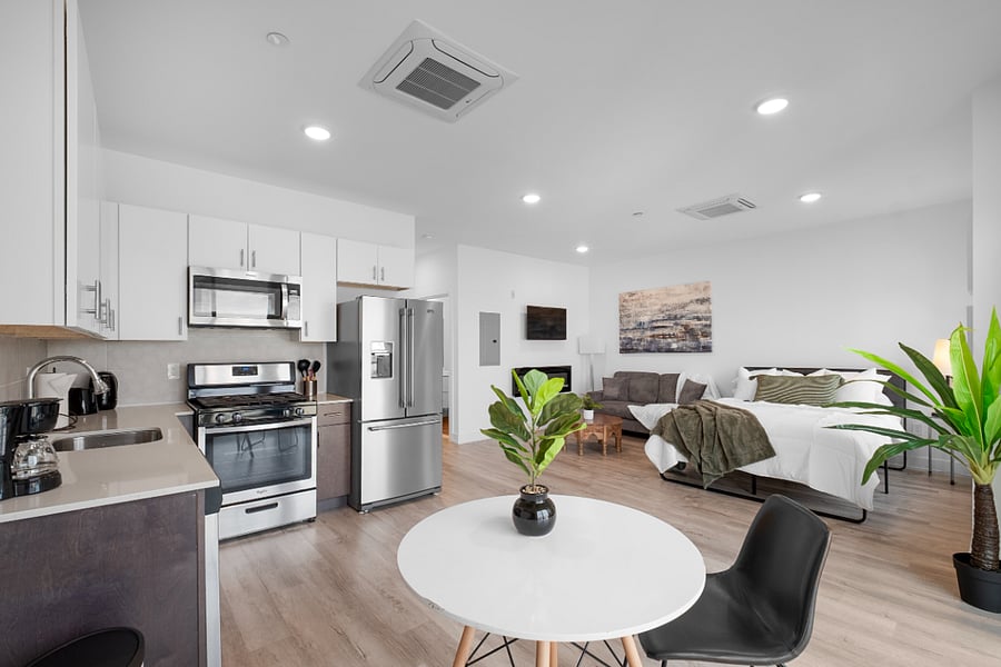 Stylish studio apartment with an open-concept layout. A neatly made bed with crisp white linens and blue accent pillows anchors the space, while a cozy sitting area with blue armchairs and a woven ottoman adds charm. The modern kitchen features stainless steel appliances, white cabinetry, and a sleek gray countertop with two black bar stools. A wall-mounted TV and electric fireplace complete the contemporary look, all set against light wood flooring and clean white walls.