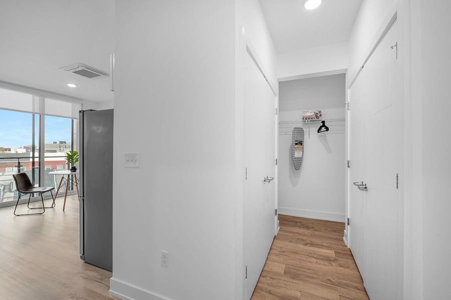 Sleek, modern apartment interior with a bright hallway featuring white walls and light wood flooring. Showcasing a stainless steel refrigerator, minimalist furniture, and large windows that flood the space with natural light