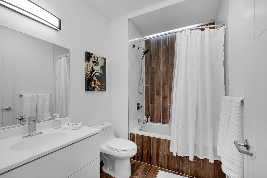 Sleek, modern bathroom with a minimalist aesthetic. It features a bathtub with a glass panel and wood-patterned tile accent wall, complemented by matching floor tiles. A white toilet and vanity with a built-in sink sit beneath a large mirror and a black light fixture. The clean white walls and subtle touches, like the purple waste bin, create a fresh and contemporary feel