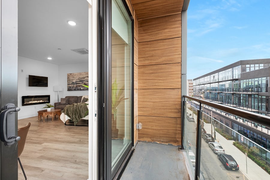 a stylish urban apartment view from a balcony with glass railing and wood-paneled walls. Through the sliding glass door, a cozy living room is visible, featuring a mounted TV, wall-mounted fireplace, wooden coffee table, and a green throw on the couch.