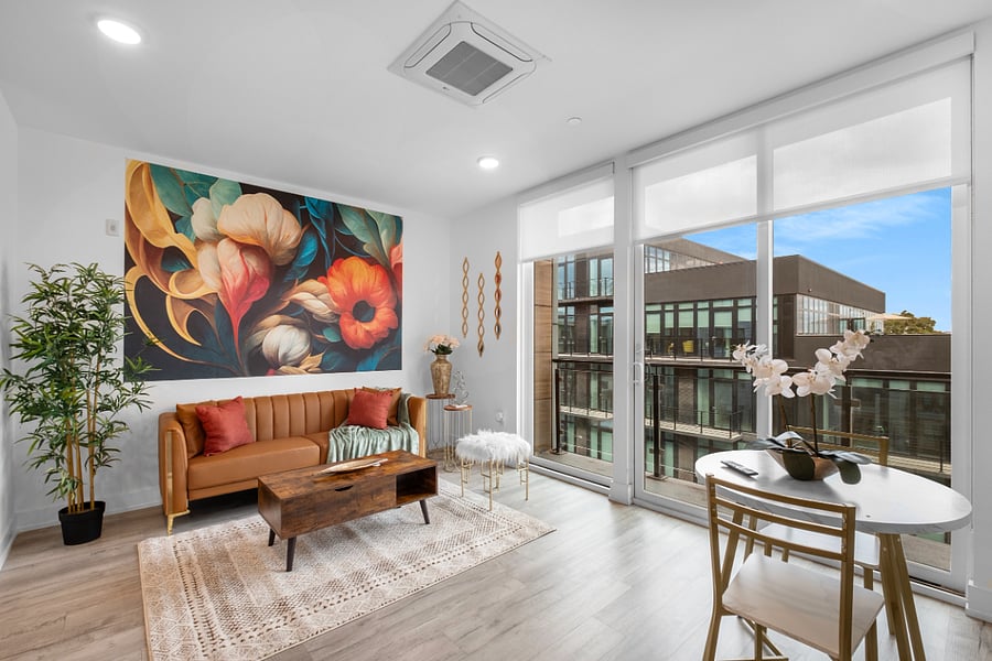 a bright, modern living room with floor-to-ceiling windows that offer a view of sleek urban architecture. Inside, a tan leather sofa with colorful pillows sits beneath a bold floral painting. 