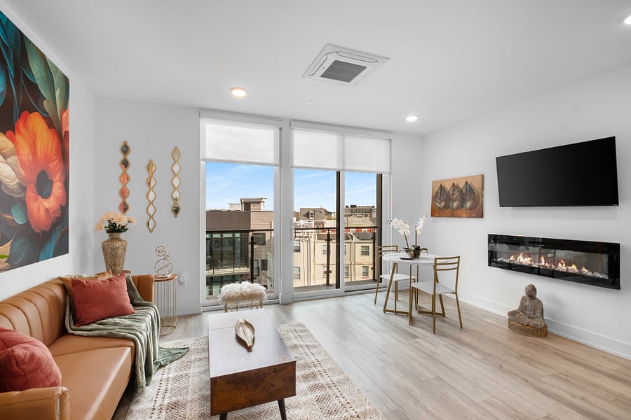 a bright, modern living room with floor-to-ceiling windows that offer a view of sleek urban architecture. Inside, a tan leather sofa with colorful pillows sits beneath a bold floral painting. 