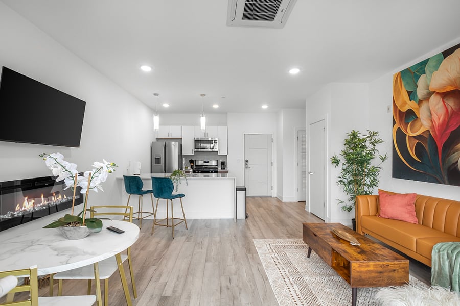 a stylish open-concept apartment with a modern kitchen and cozy living area. The kitchen features a white island with a built-in sink and dishwasher, dark brown cabinets, and pendant lighting. Adjacent to the kitchen, the living space includes a tan leather sofa with colorful pillows, a wooden coffee table, and a vibrant floral wall art.