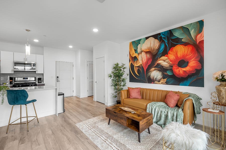a stylish open-concept apartment with a modern kitchen and cozy living area. The kitchen features a white island with a built-in sink and dishwasher, dark brown cabinets, and pendant lighting. Adjacent to the kitchen, the living space includes a tan leather sofa with colorful pillows, a wooden coffee table, and a vibrant floral wall art.