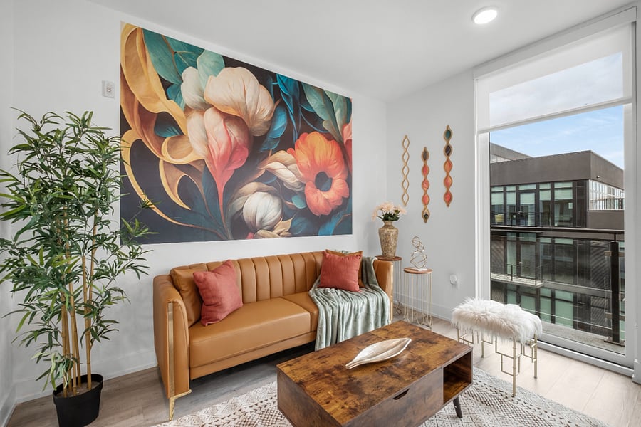 a bright, modern living room with floor-to-ceiling windows that offer a view of sleek urban architecture. Inside, a tan leather sofa with colorful pillows sits beneath a bold floral painting. 