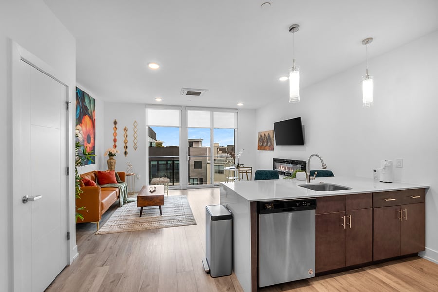 a stylish open-concept apartment with a modern kitchen and cozy living area. The kitchen features a white island with a built-in sink and dishwasher, dark brown cabinets, and pendant lighting. Adjacent to the kitchen, the living space includes a tan leather sofa with colorful pillows, a wooden coffee table, and a vibrant floral wall art.