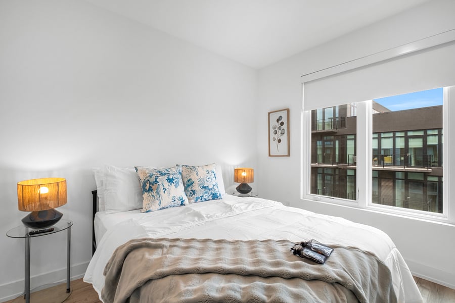 Minimalist bedroom with white walls and light wood flooring, featuring a large bed dressed in white linens and blue accent pillows. Warm bedside lamps sit on round tables, and a soft throw adds texture. A wall-mounted TV faces the bed, while a large window offers natural light and a view of a modern building.
