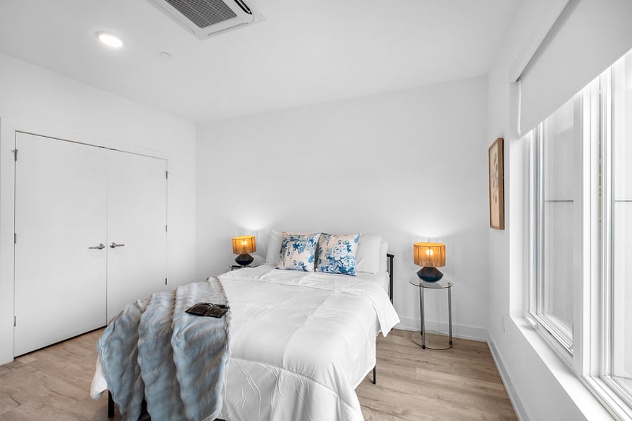 Bright, minimalist bedroom with white walls and light wood flooring, featuring a neatly made bed with white linens and blue floral accent pillows. An open closet with double doors reveals shelving and a woven decor piece.