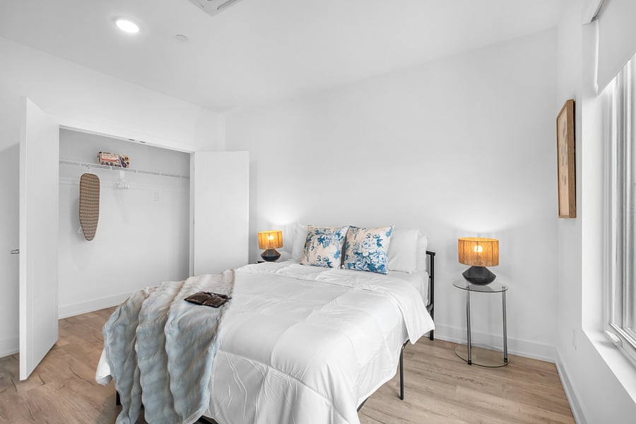 Bright, minimalist bedroom with white walls and light wood flooring, featuring a neatly made bed with white linens and blue floral accent pillows. An open closet with double doors reveals shelving and a woven decor piece.