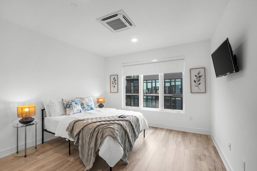 Minimalist bedroom with white walls and light wood flooring, featuring a large bed dressed in white linens and blue accent pillows. Warm bedside lamps sit on round tables, and a soft throw adds texture. A wall-mounted TV faces the bed, while a large window offers natural light and a view of a modern building.
