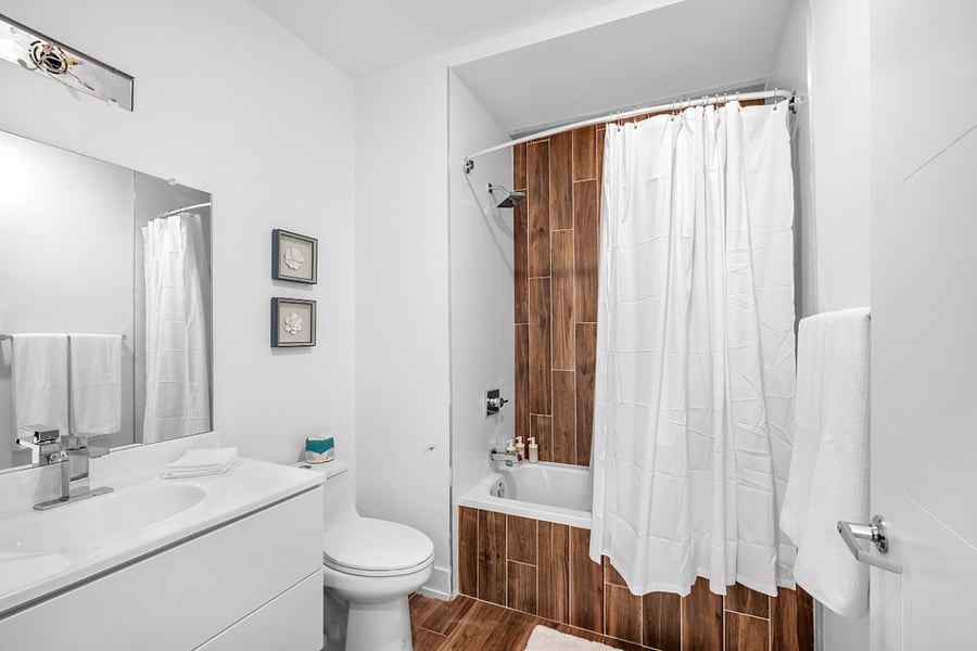 Sleek, modern bathroom with a minimalist aesthetic. It features a bathtub with a glass panel and wood-patterned tile accent wall, complemented by matching floor tiles. A white toilet and vanity with a built-in sink sit beneath a large mirror and a black light fixture. The clean white walls and subtle touches, like the purple waste bin, create a fresh and contemporary feel