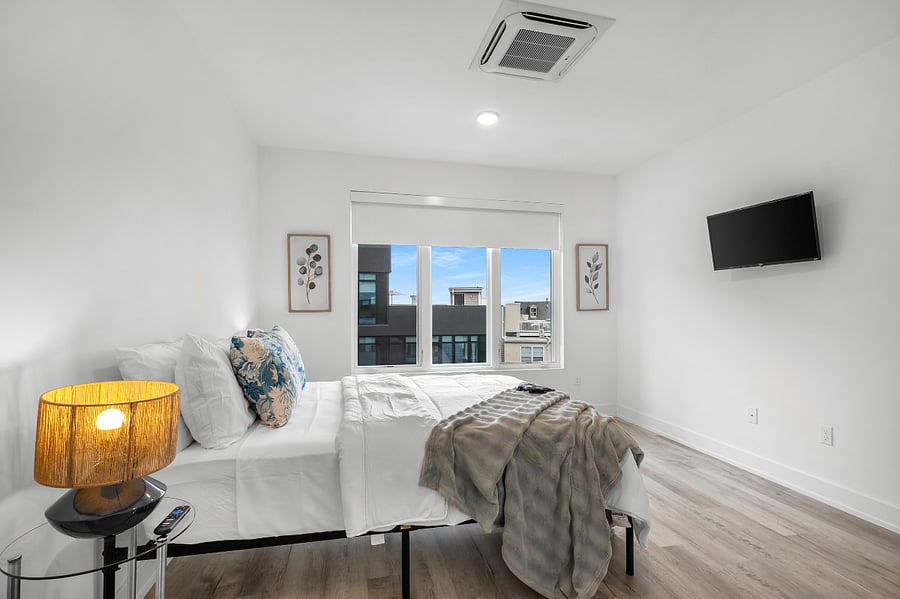 Minimalist bedroom with white walls and light wood flooring, featuring a large bed dressed in white linens and blue accent pillows. Warm bedside lamps sit on round tables, and a soft throw adds texture. A wall-mounted TV faces the bed, while a large window offers natural light and a view of a modern building.
