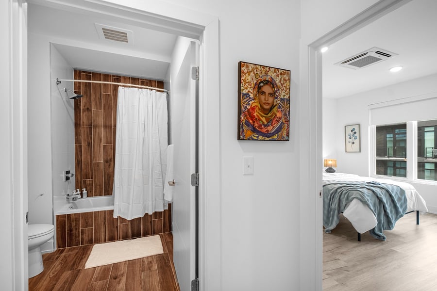 A bright, modern apartment interior featuring a bathroom and adjacent bedroom. The bathroom has warm wood-look tile, a white bathtub with a shower curtain, and a sleek toilet.