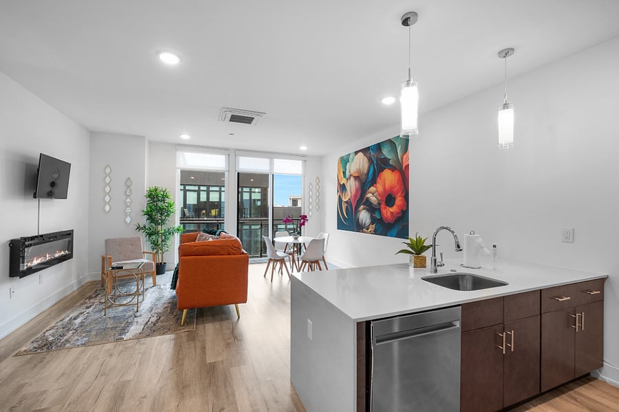 a stylish open-concept apartment with a modern kitchen and cozy living area. The kitchen features a white island with a built-in sink and dishwasher, dark brown cabinets, and pendant lighting. Adjacent to the kitchen, the living space includes a tan leather sofa with colorful pillows, a wooden coffee table, and a vibrant floral wall art.