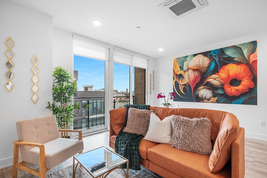 a bright, modern living room with floor-to-ceiling windows that offer a view of sleek urban architecture. Inside, a tan leather sofa with colorful pillows sits beneath a bold floral painting. 
