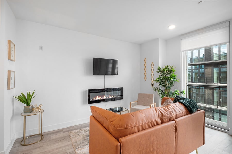 a bright, modern living room with floor-to-ceiling windows that offer a view of sleek urban architecture. Inside, a tan leather sofa with colorful pillows sits beneath a bold floral painting. 