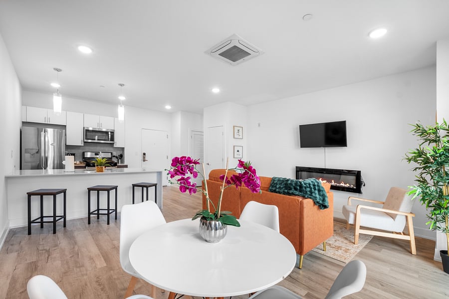 a stylish open-concept apartment with a modern kitchen and cozy living area. The kitchen features a white island with a built-in sink and dishwasher, dark brown cabinets, and pendant lighting. Adjacent to the kitchen, the living space includes a tan leather sofa with colorful pillows, a wooden coffee table, and a vibrant floral wall art.
