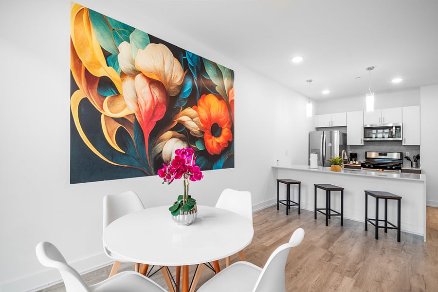 a stylish open-concept apartment with a modern kitchen and cozy living area. The kitchen features a white island with a built-in sink and dishwasher, dark brown cabinets, and pendant lighting. Adjacent to the kitchen, the living space includes a tan leather sofa with colorful pillows, a wooden coffee table, and a vibrant floral wall art.