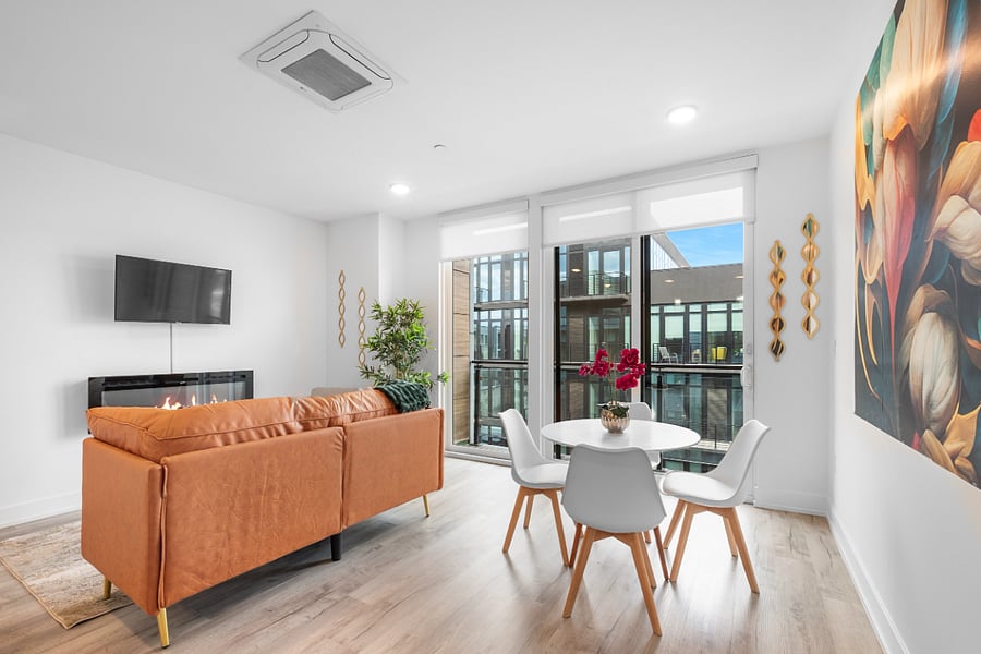 a bright, modern living room with floor-to-ceiling windows that offer a view of sleek urban architecture. Inside, a tan leather sofa with colorful pillows sits beneath a bold floral painting. 