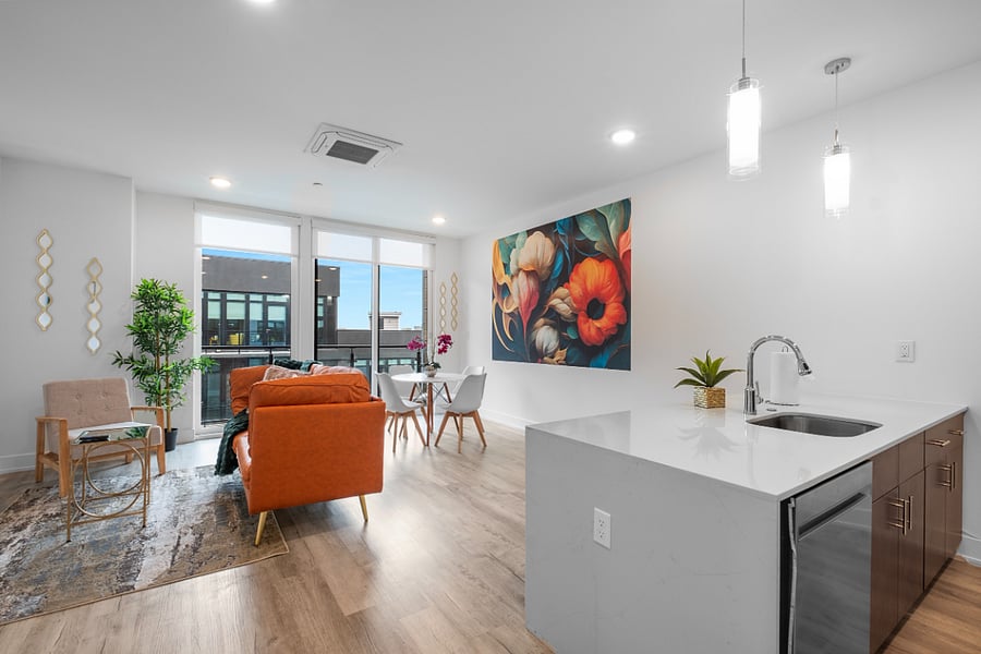 a bright, modern living room with floor-to-ceiling windows that offer a view of sleek urban architecture. Inside, a tan leather sofa with colorful pillows sits beneath a bold floral painting. 