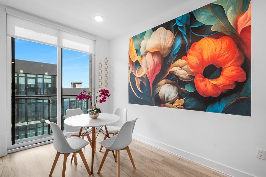 a bright, modern living room with floor-to-ceiling windows that offer a view of sleek urban architecture. Inside, a tan leather sofa with colorful pillows sits beneath a bold floral painting. 