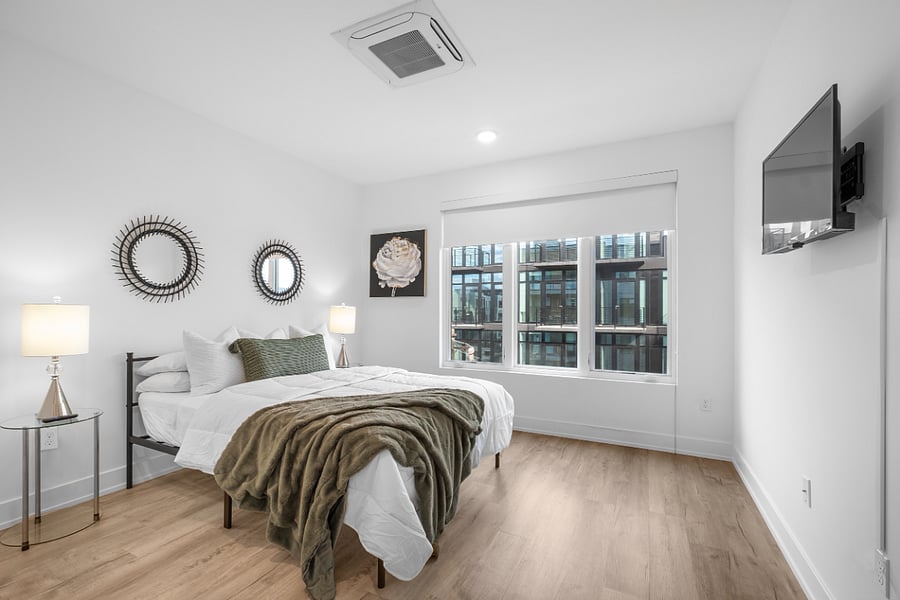 Modern bedroom with white walls and light wood flooring, featuring a neatly made bed with white linens, green accent pillow, and matching throw blanket. Glass-top nightstands with silver lamps flank the bed, while decorative spiked mirrors and floral artwork add visual interest. A large window provides natural light and a view of a contemporary building exterior.
