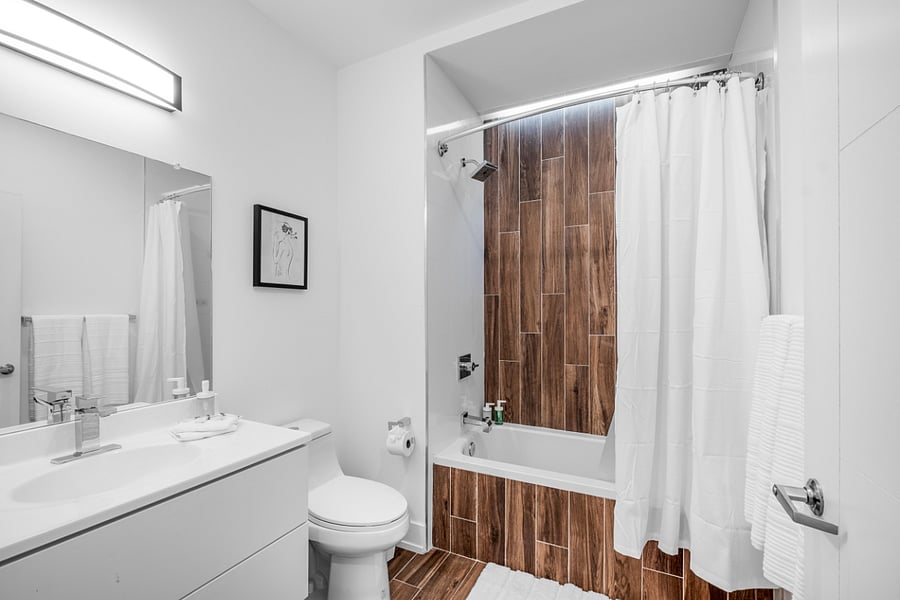Sleek, modern bathroom with a minimalist aesthetic. It features a bathtub with a glass panel and wood-patterned tile accent wall, complemented by matching floor tiles. A white toilet and vanity with a built-in sink sit beneath a large mirror and a black light fixture. The clean white walls and subtle touches, like the purple waste bin, create a fresh and contemporary feel