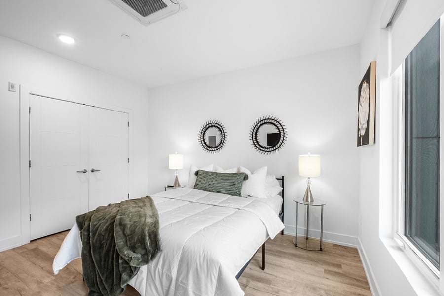 Modern bedroom with white walls and light wood flooring, featuring a neatly made bed with white linens, green accent pillow, and matching throw blanket.