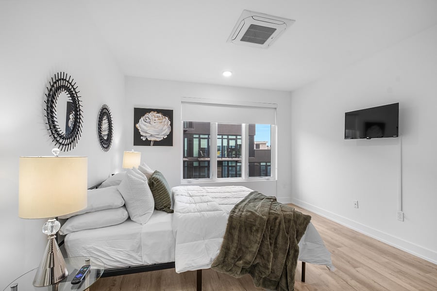 Modern bedroom with white walls and light wood flooring, featuring a neatly made bed with white linens, green accent pillow, and matching throw blanket. Glass-top nightstands with silver lamps flank the bed, while decorative spiked mirrors and floral artwork add visual interest. A large window provides natural light and a view of a contemporary building exterior.
