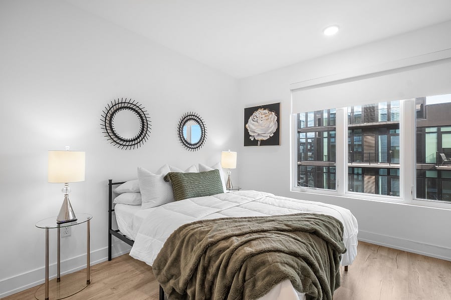 Modern bedroom with white walls and light wood flooring, featuring a neatly made bed with white linens, green accent pillow, and matching throw blanket. Glass-top nightstands with silver lamps flank the bed, while decorative spiked mirrors and floral artwork add visual interest. A large window provides natural light and a view of a contemporary building exterior.
