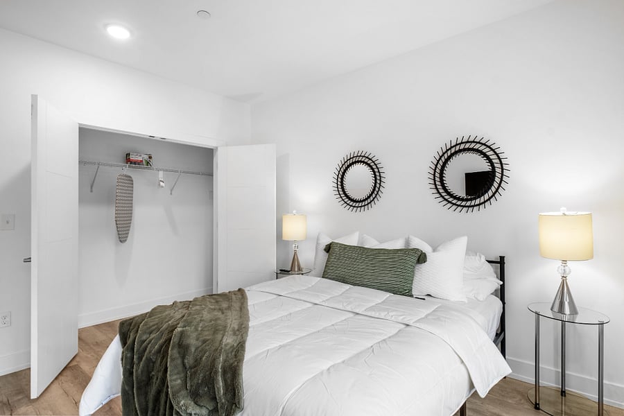 Minimalist bedroom with white walls and light wood flooring, featuring a neatly made bed with white linens, green accent pillow, and matching throw blanket.An open closet reveals shelving and a hanging ironing board.