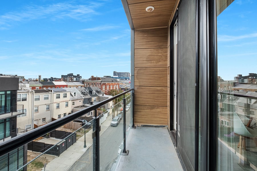 Urban balcony view from a high-rise apartment featuring a glass railing with metal supports, concrete flooring, and wood-paneled wall cladding. 