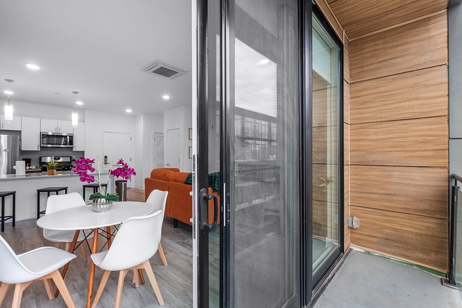 Modern apartment interior with open-plan layout featuring a white kitchen with stainless steel appliances, island seating, and a round dining table with white chairs and vibrant purple orchids. The living area includes an orange sofa with a green throw. Glass sliding doors lead to a balcony with wood-paneled walls, enhancing the contemporary design. Light gray wood flooring and white walls unify the space.
